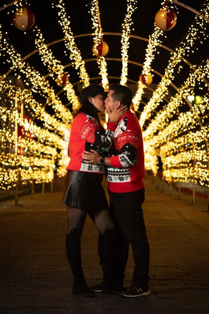 pexels-photo-29897459-29897459 Couple in red sweaters kissing under illuminated Christmas decorations at night.