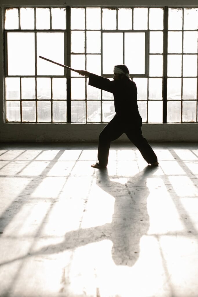 Silhouette of martial artist practicing with a staff in a sunlit studio.