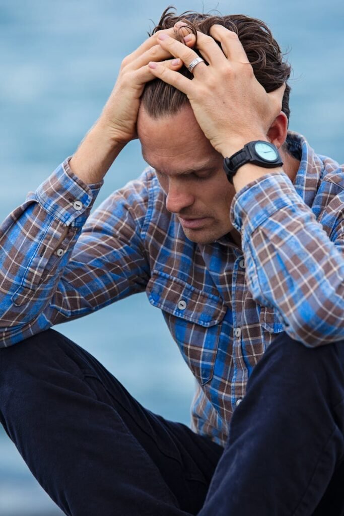 A man in a plaid shirt sits by the water looking distressed, symbolizing stress.