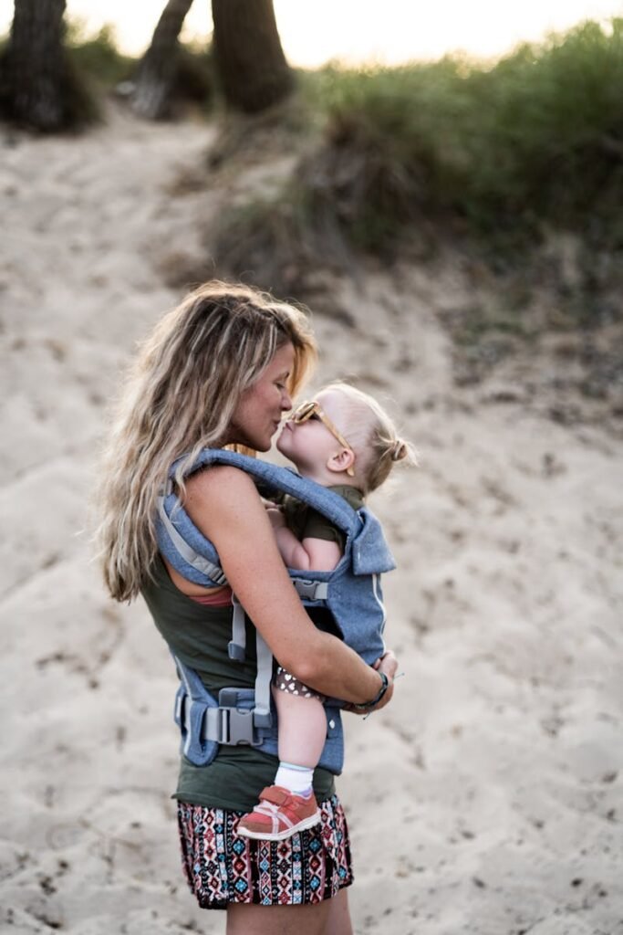 pexels-photo-2701598-2701598 A loving mother kisses her toddler in a baby carrier on a sandy beach, capturing a tender parenting moment.